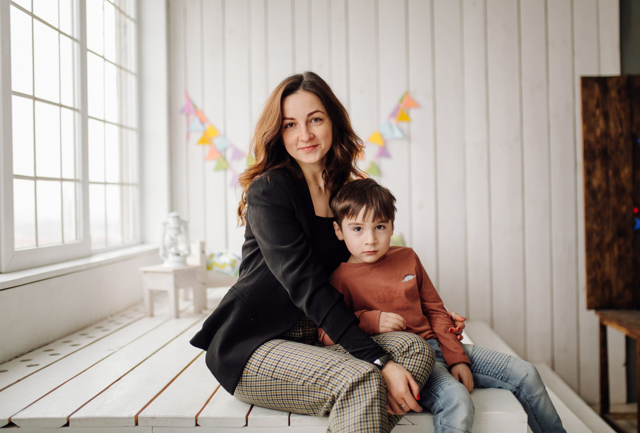 Mother and son posing together in a studio setting, wearing casual clothes, with colorful decorations in the background, representing family and care related to Kids R Kids franchise.