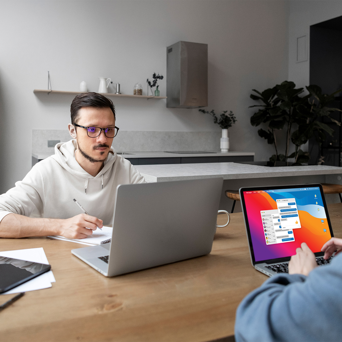 Person in a hoodie working on a laptop at a table, with another laptop displaying a communication interface, in a modern workspace environment.