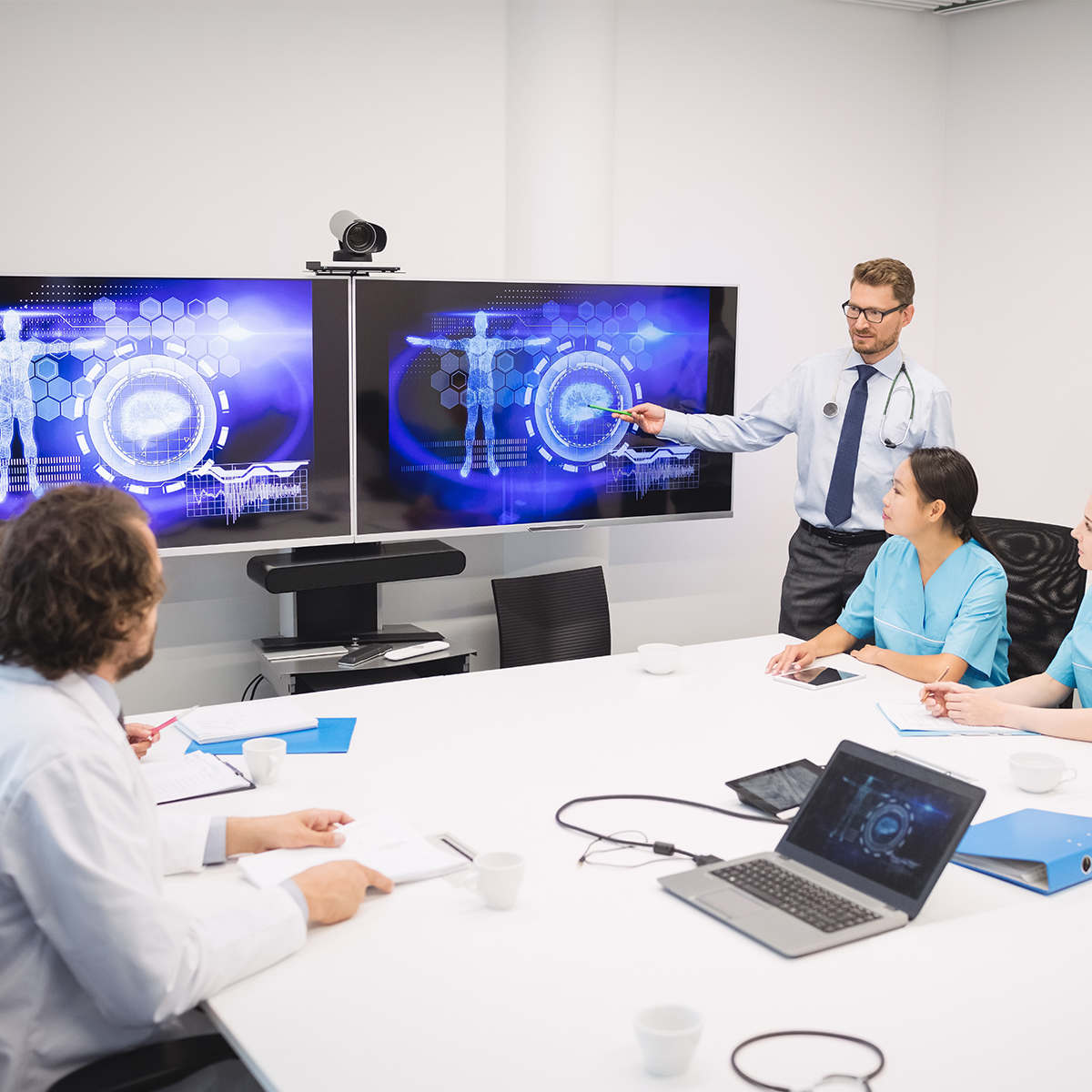 Healthcare professionals in a meeting room discussing digital health technology on dual monitors, with a doctor presenting data visualizations related to patient care and EHR systems, while colleagues take notes and engage in the discussion.